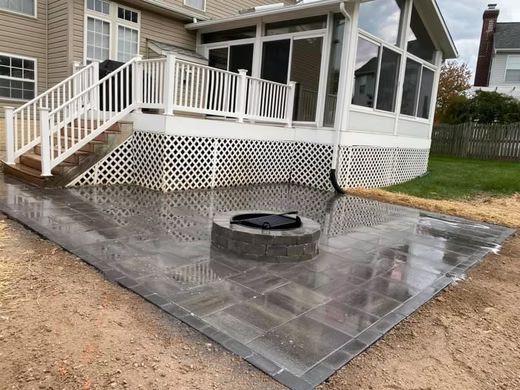 Patio with gray pavers and circular fire pit beside a house with white railings and lattice skirting