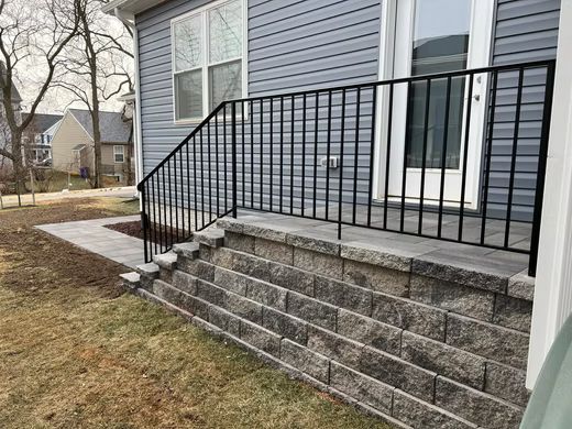 Front steps with black metal railing leading to a gray house entrance over a stone retaining wall.