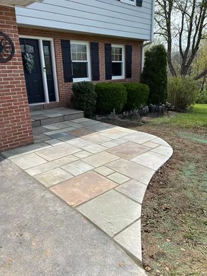 Curved stone walkway beside a brick house and front steps, bordered by shrubs and lawn