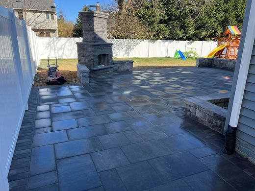 Patio with dark stone pavers, outdoor brick grill, and playset in a backyard surrounded by a white fence