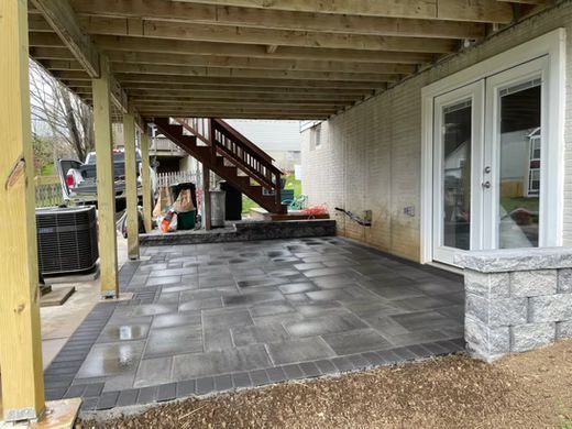 Covered patio with gray paver flooring, wooden stairs, and white French doors beside a stone foundation.