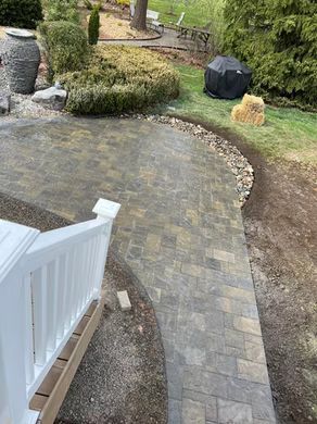 Curved stone patio and walkway beside a white railing, with bushes and a covered grill in a backyard