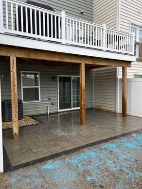 Covered backyard patio under a white-railed deck with gray siding and wet stone flooring