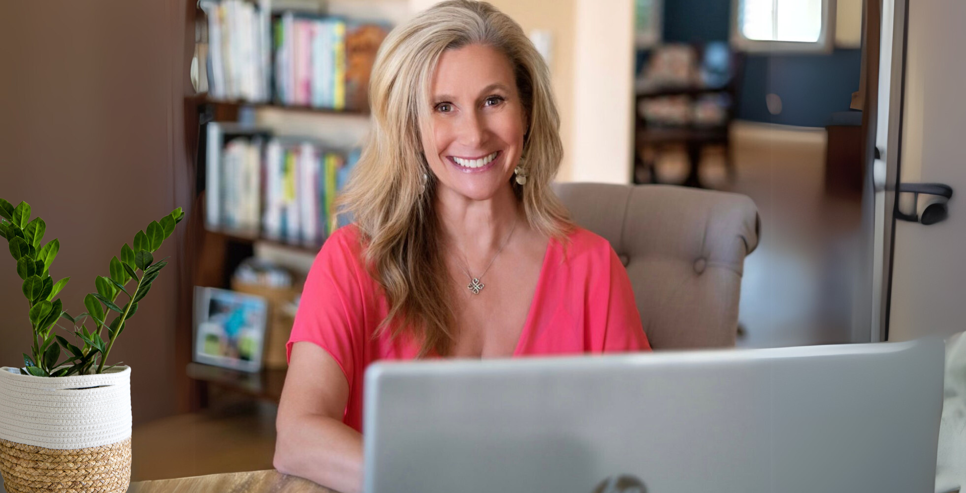 A woman with blonde hair, smiling at a laptop, wearing a pink top, in a home office with a bookshelf.