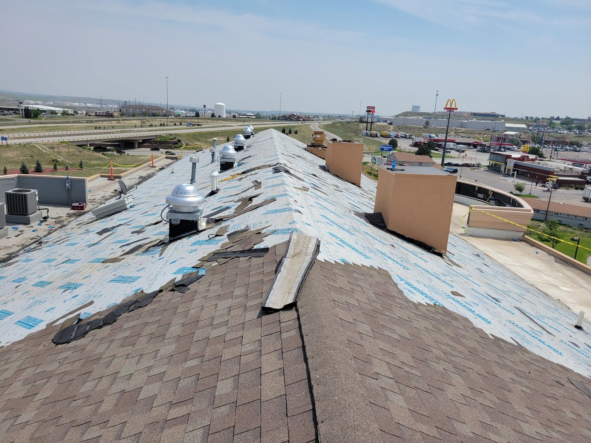 Rooftop under repair with torn blue underlayment, shingles, and rooftop vents overlooking a cityscape