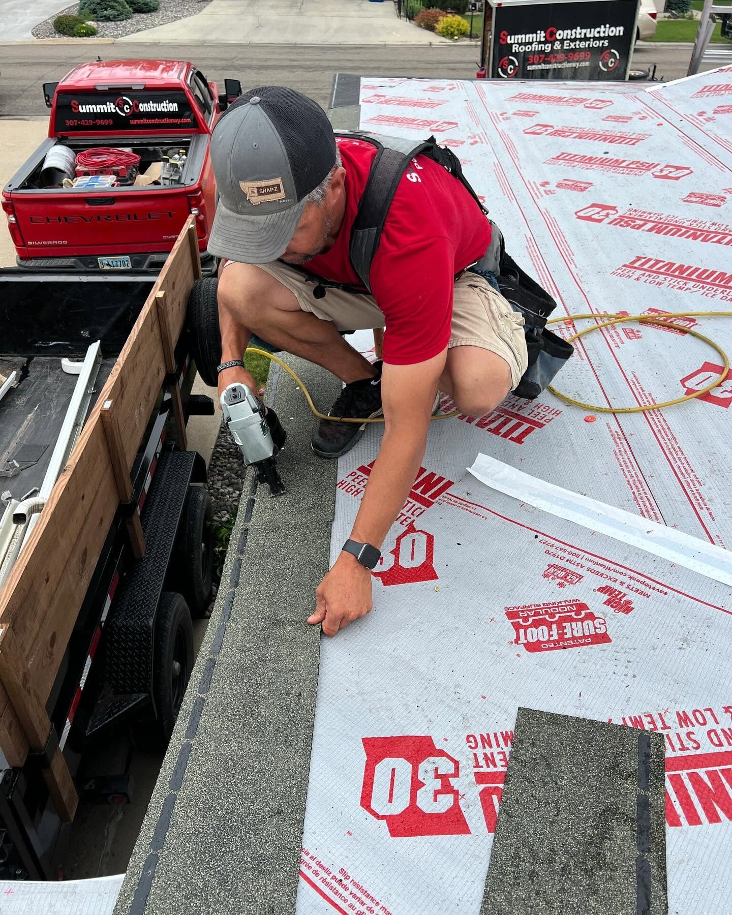 Worker kneeling on rooftop, fastening roofing underlayment beside a truck bed of supplies.