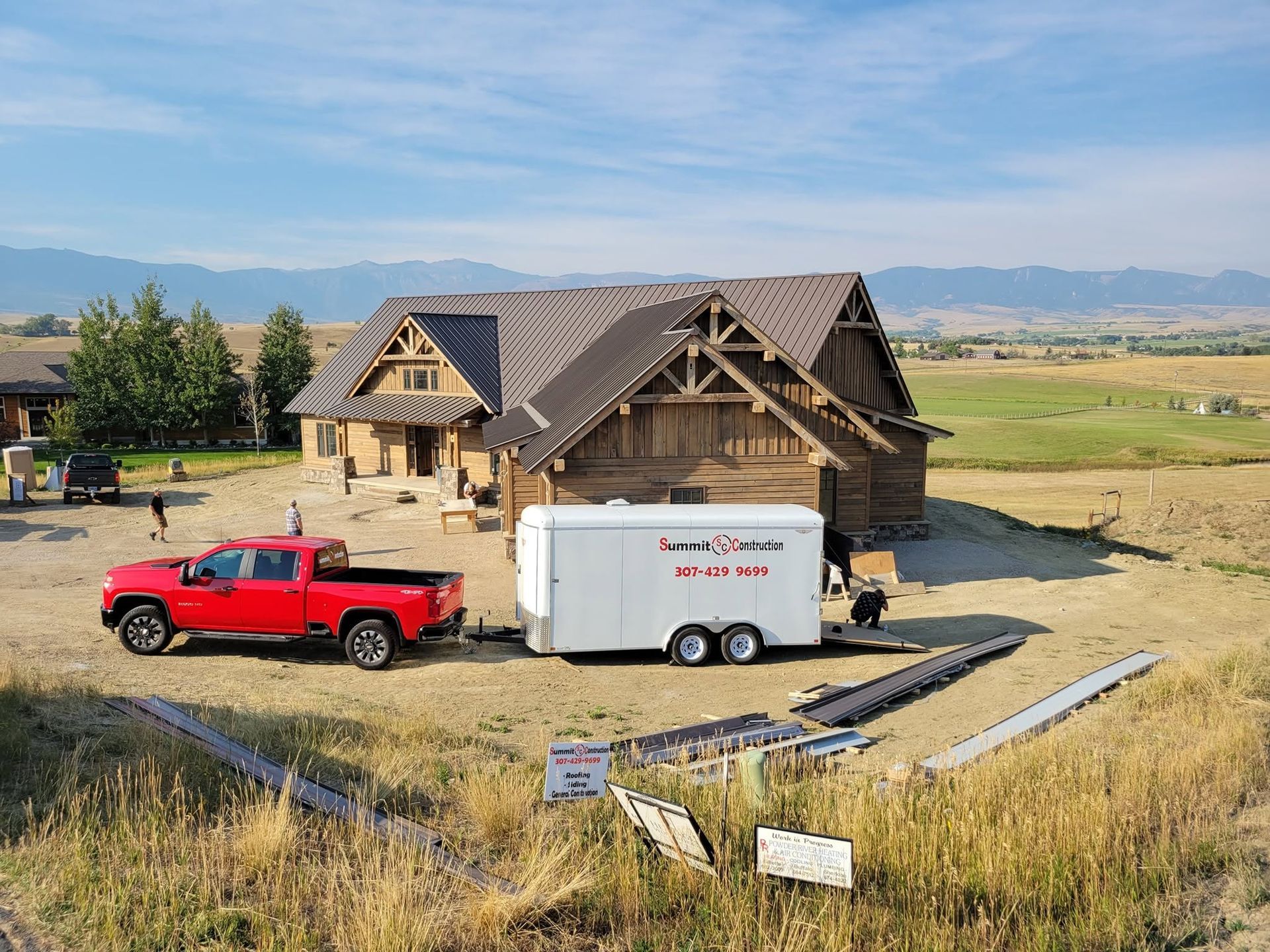Red pickup truck and white trailer parked beside a partially built house on a grassy lot with mountains in the distance