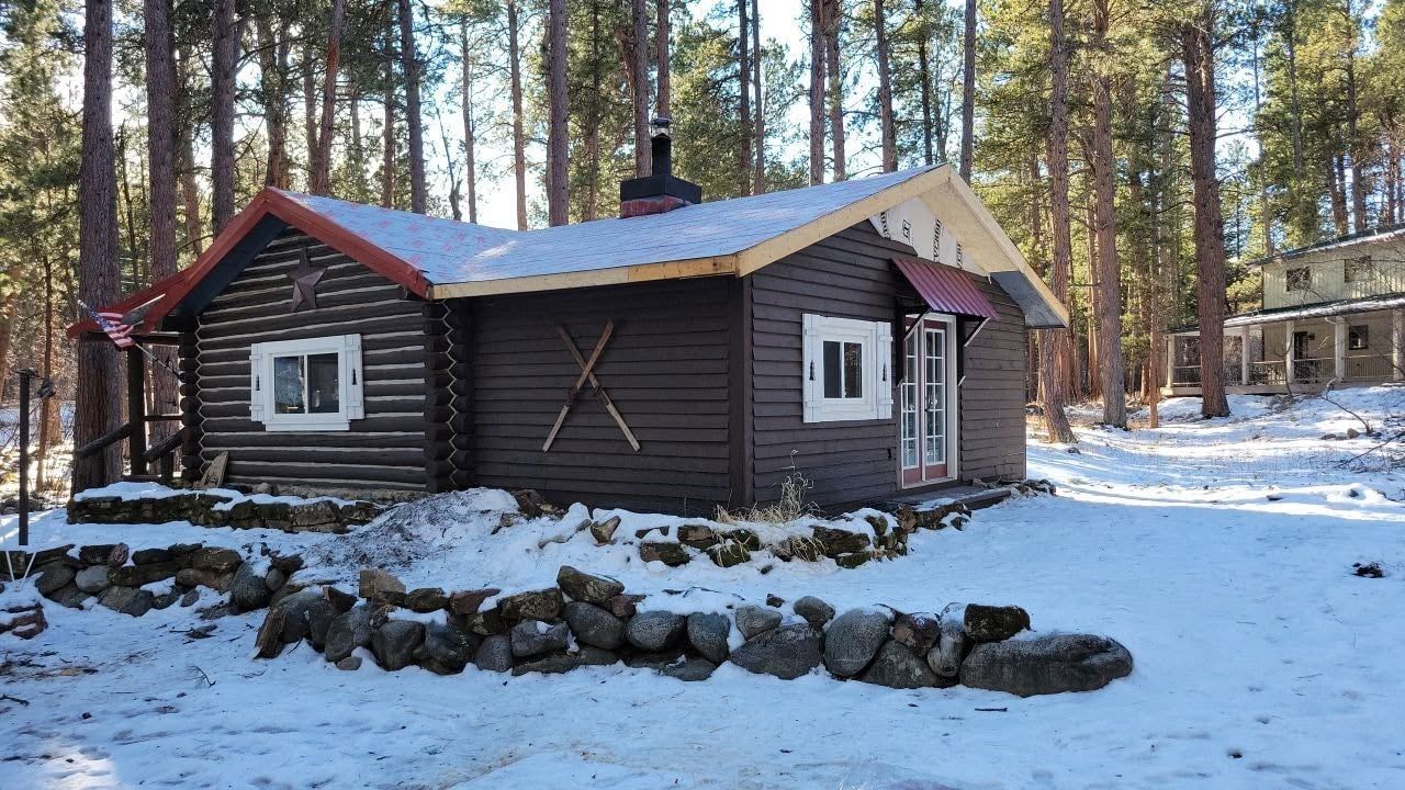 Snowy cabin in a pine forest, with a stone border and a smaller building in the background.