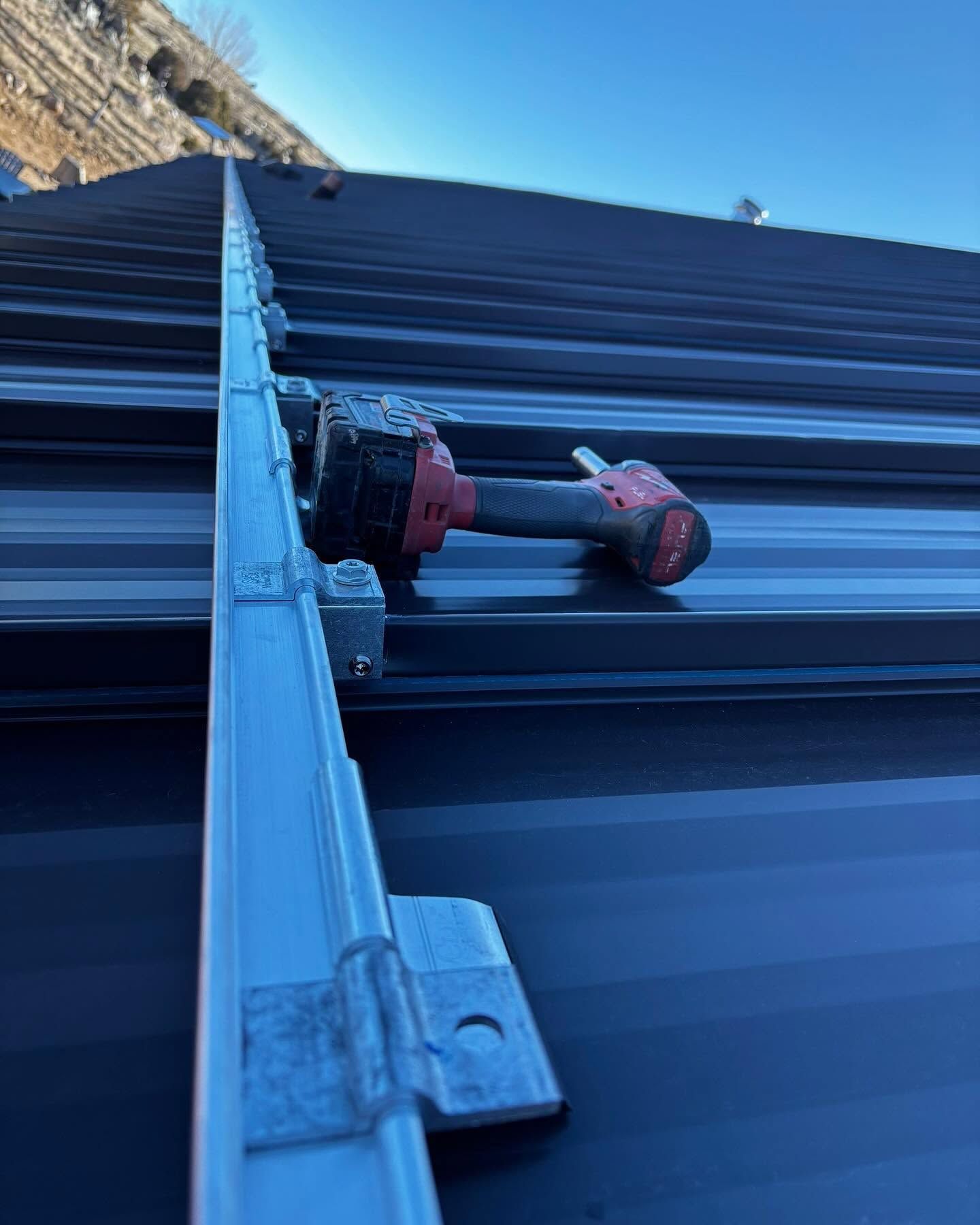 Drill-mounted roof bracket aligned on dark corrugated metal roofing under a blue sky