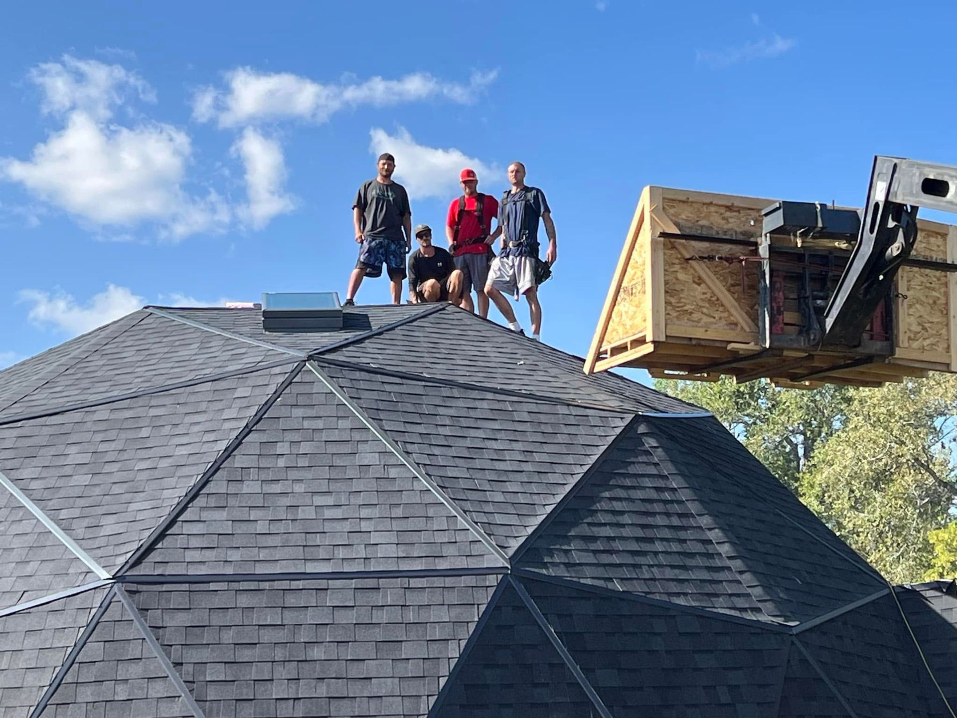 Three people on a shingled roof beside a lifted bucket truck under a blue sky