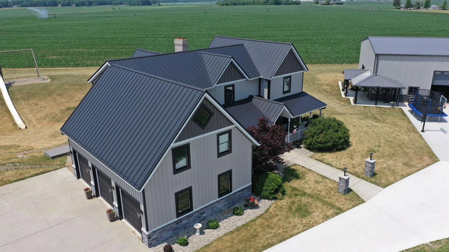 Aerial view of a gray suburban house with black roof, driveway, and lawn beside open farmland