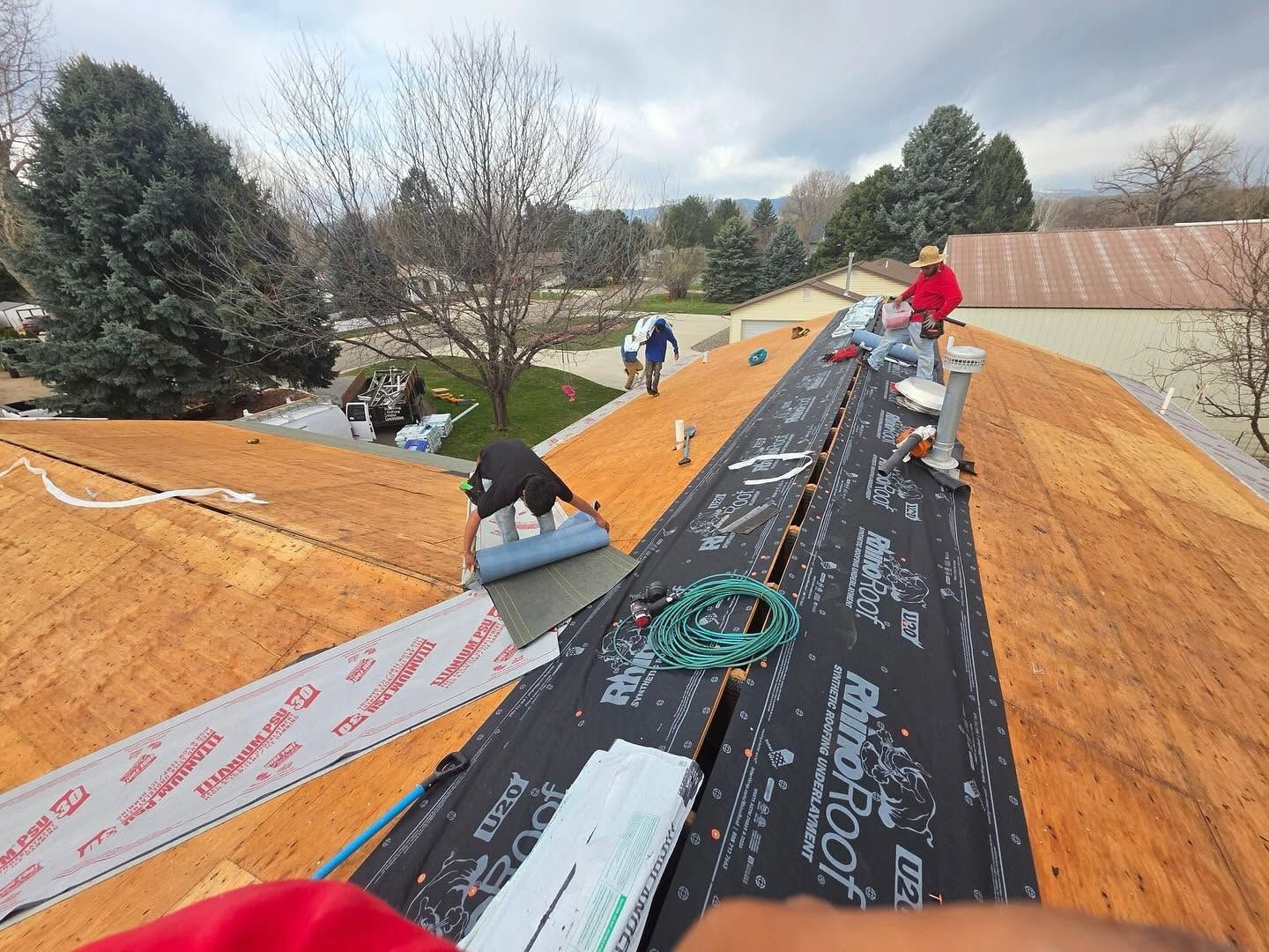 Workers installing roofing underlayment on a steep orange roof, with tools and trees in the background.
