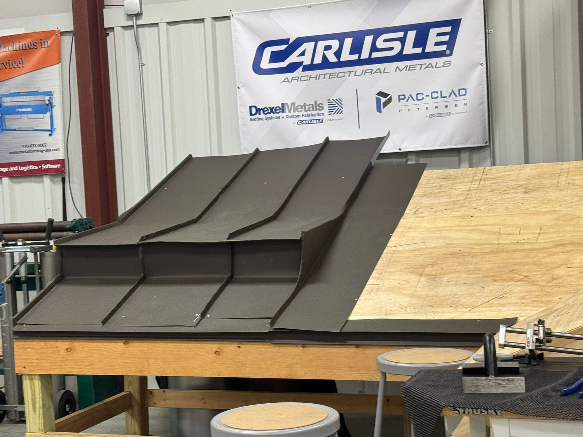 Black metal roofing panels on a workbench in a workshop, with a Carlisle banner in the background