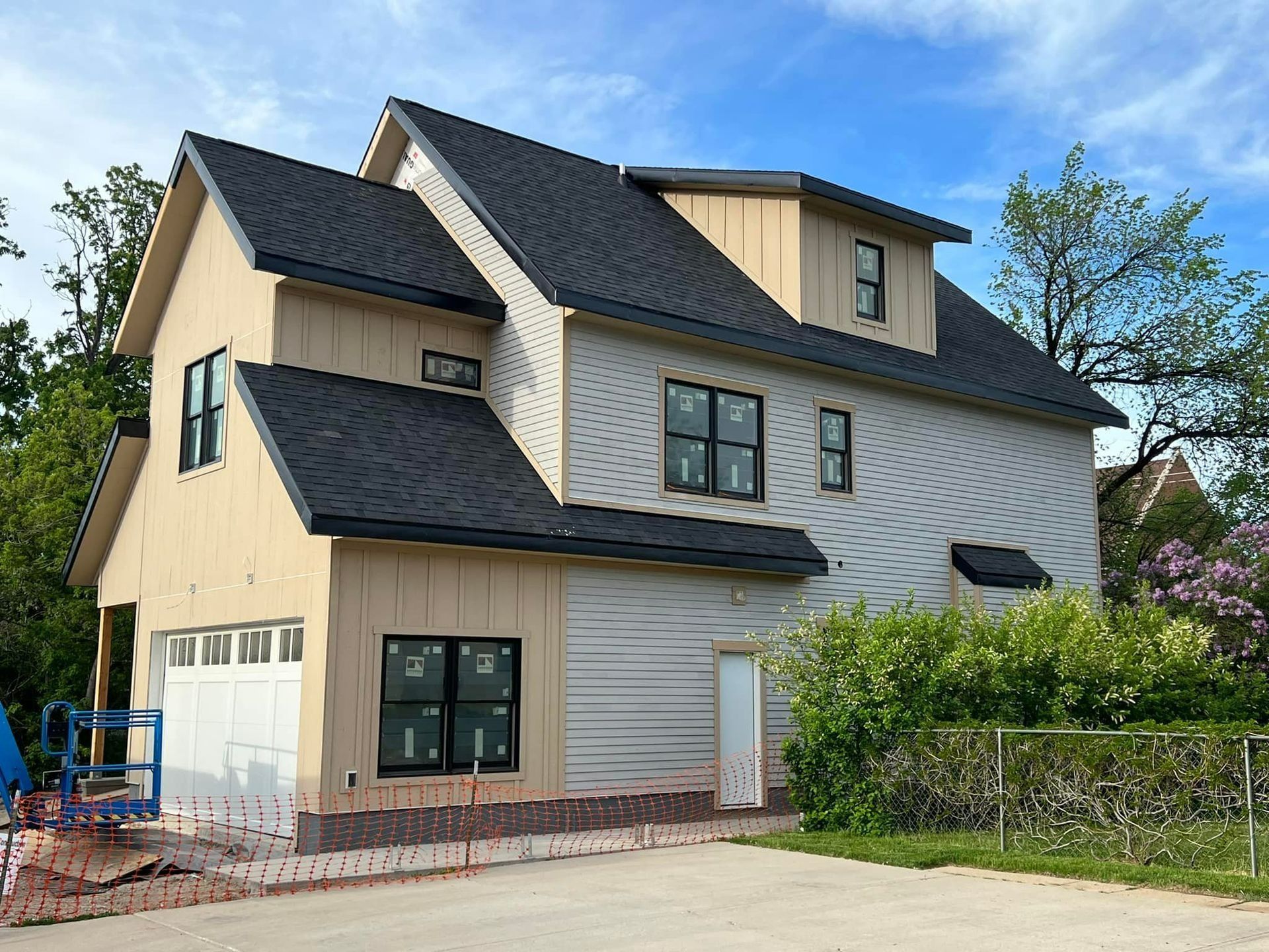 Two-story house under construction with beige siding, black roof, and a garage, set beside a fenced yard.
