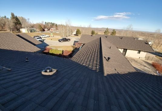 Rooftop view of dark shingle roofs, vents, and a suburban neighborhood under a clear blue sky.