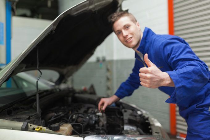 Mechanic in blue coveralls inspecting a car engine with the hood open and giving a thumbs-up