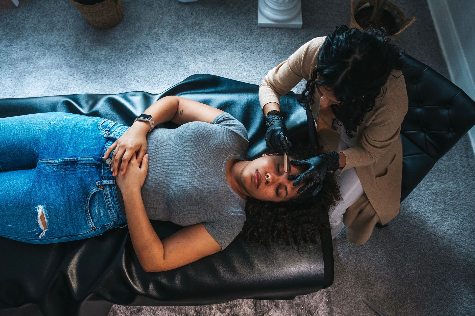 A woman is laying on a couch getting her eyebrows done by a woman.