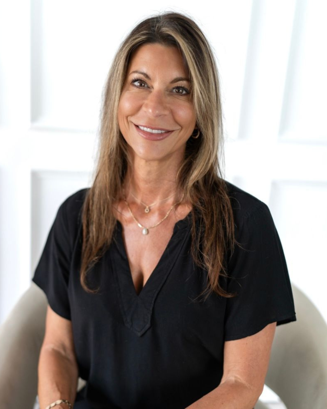 Woman with long brown hair, smiling, wearing a black shirt, seated in a chair.