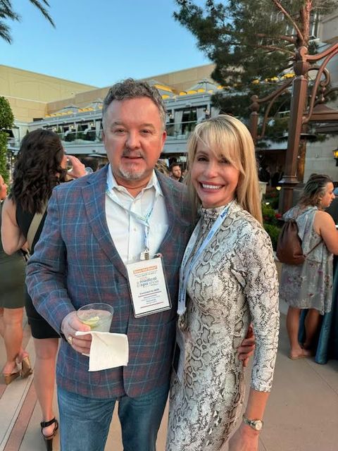 Man and woman pose together outdoors. Man in plaid jacket holds a drink, woman in snakeskin dress.