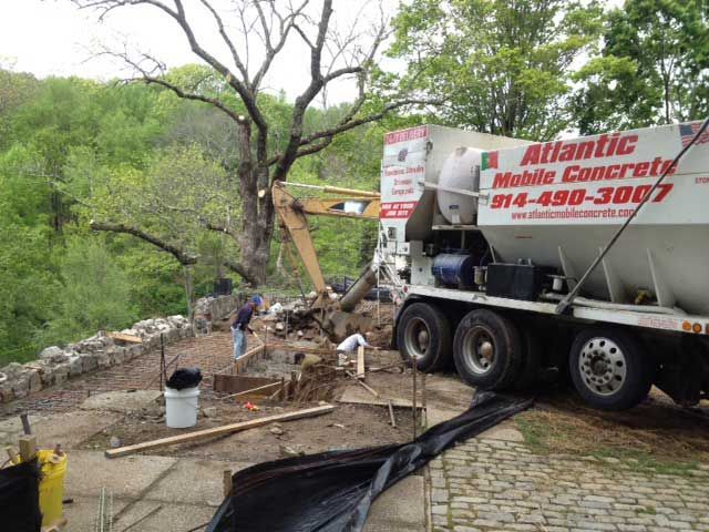 Waste removal truck at a muddy construction site beside a stone wall and trees
