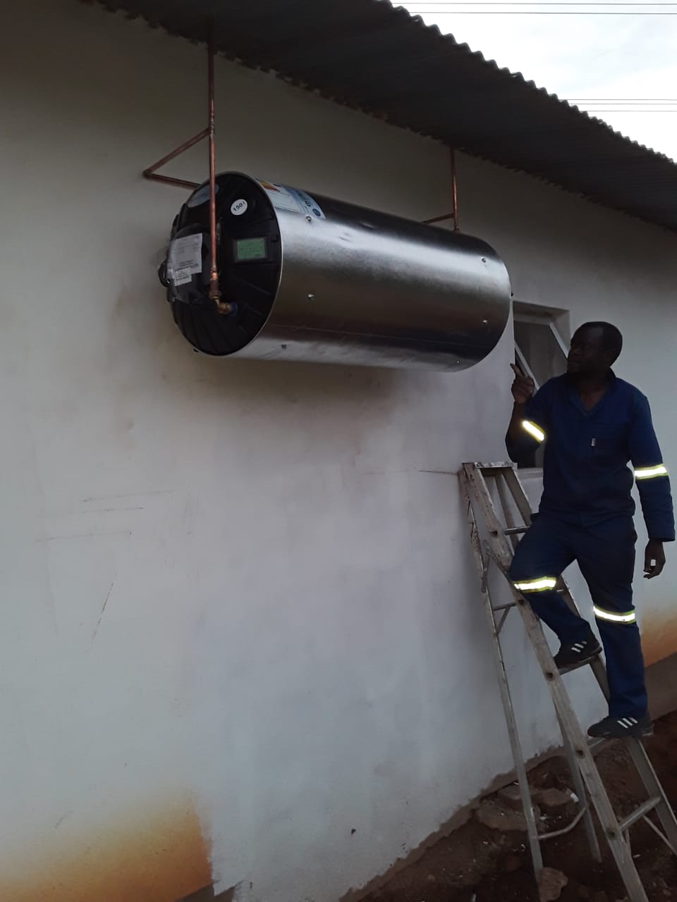 worker standing on a ladder and inspecting a wall mounted electric geyser
