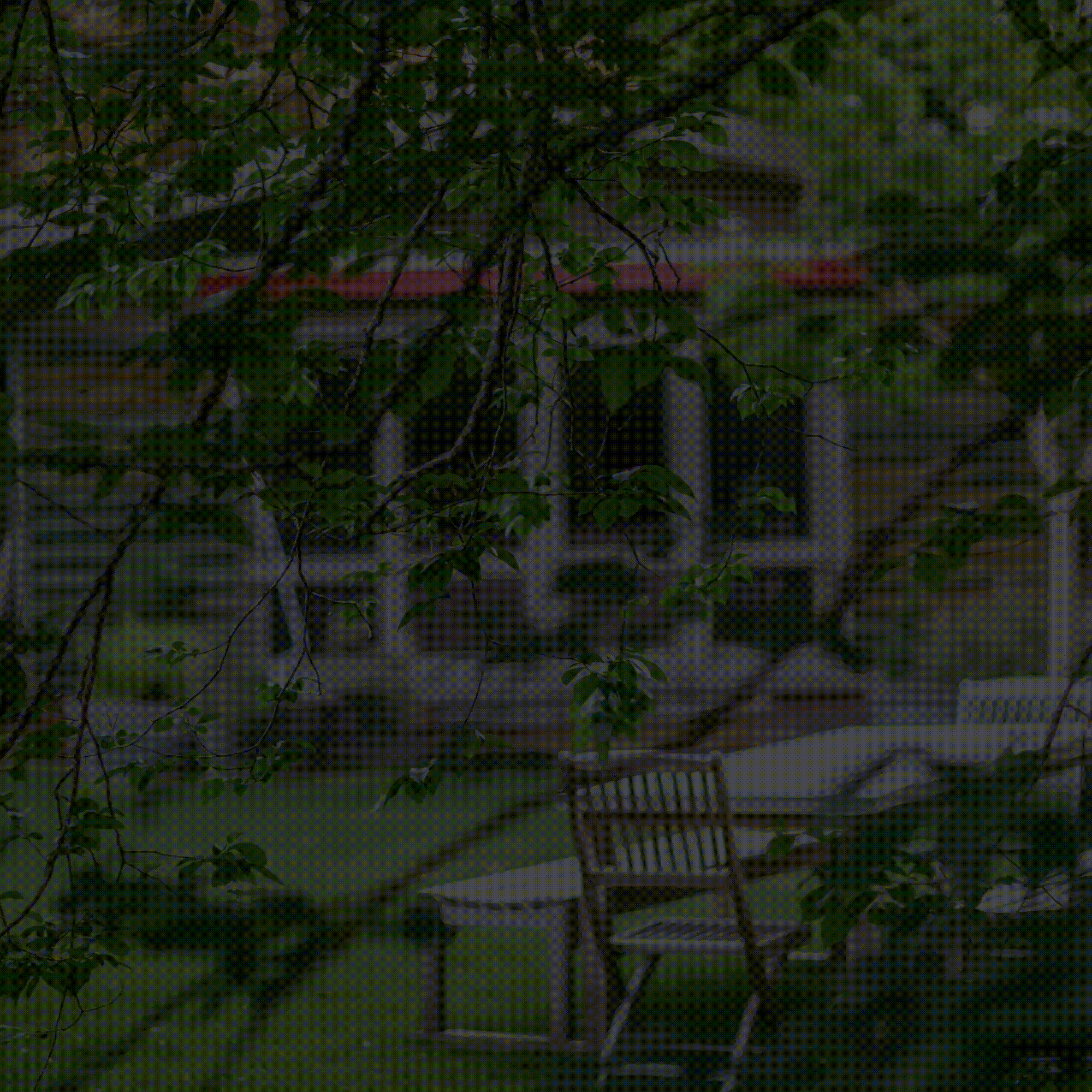 Wooden chair and table on a shaded patio beside a house, framed by leafy branches.
