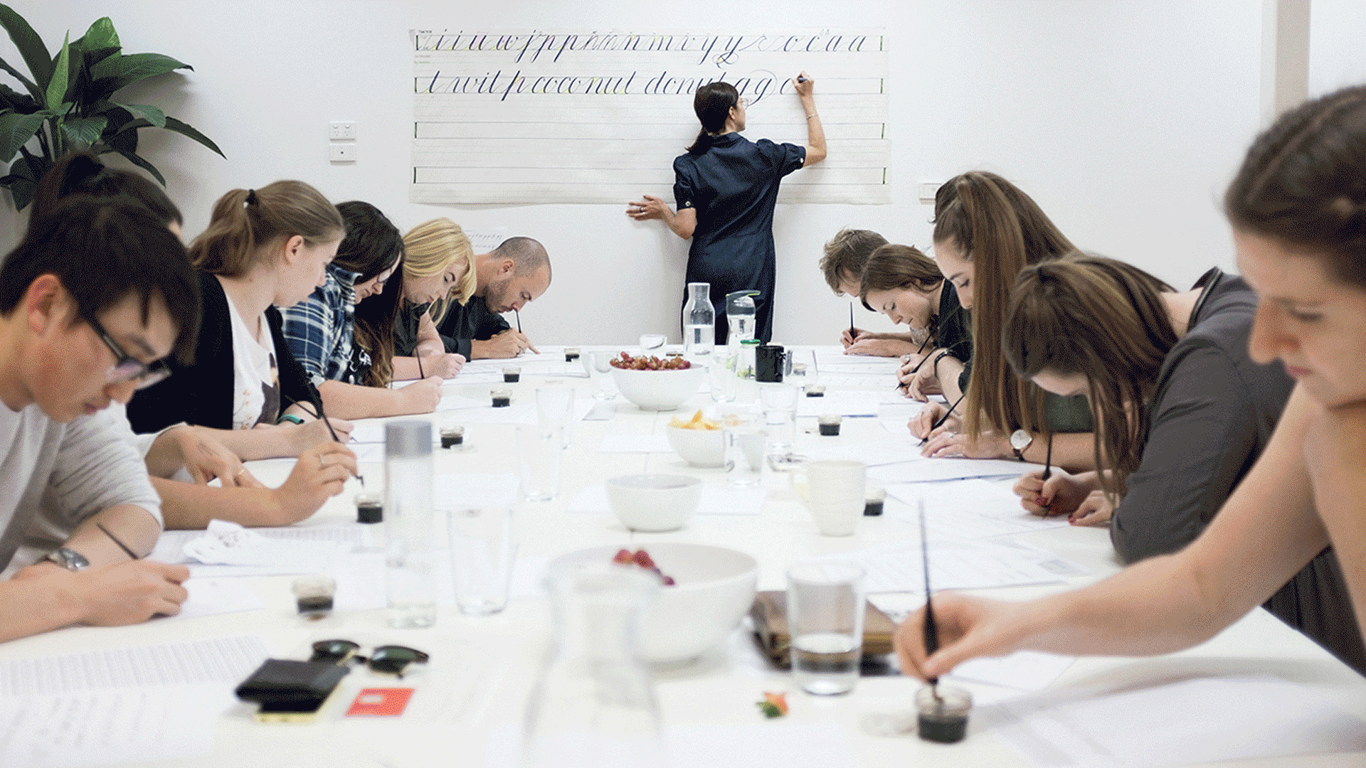 People seated around a conference table, taking notes while a presenter writes on a whiteboard.