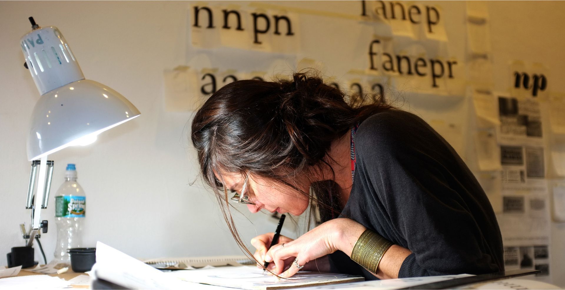 Maria Montes leaning over a desk and writing under a lamp, with letters posted on the wall behind them