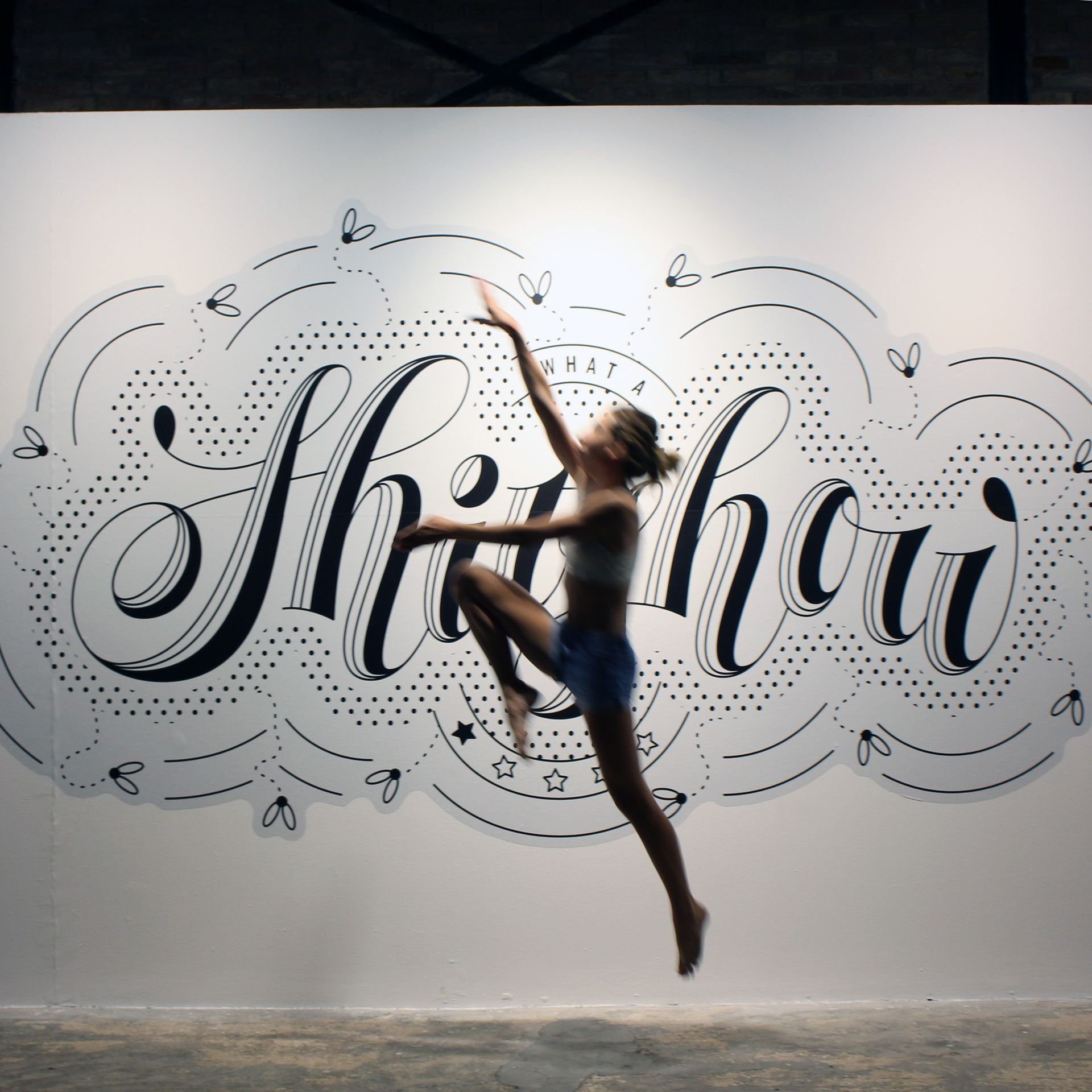 Dancer leaping in front of a white wall with large black script mural and dotted decorative swirls
