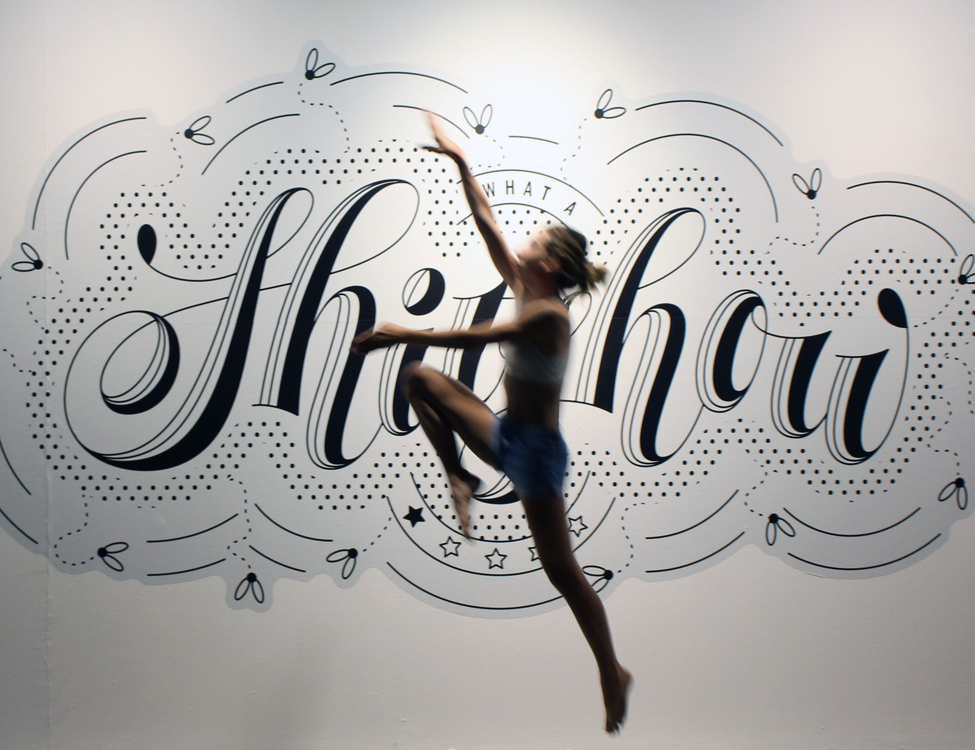A person mid-leap in front of a white wall featuring the decorative, hand-lettered phrase