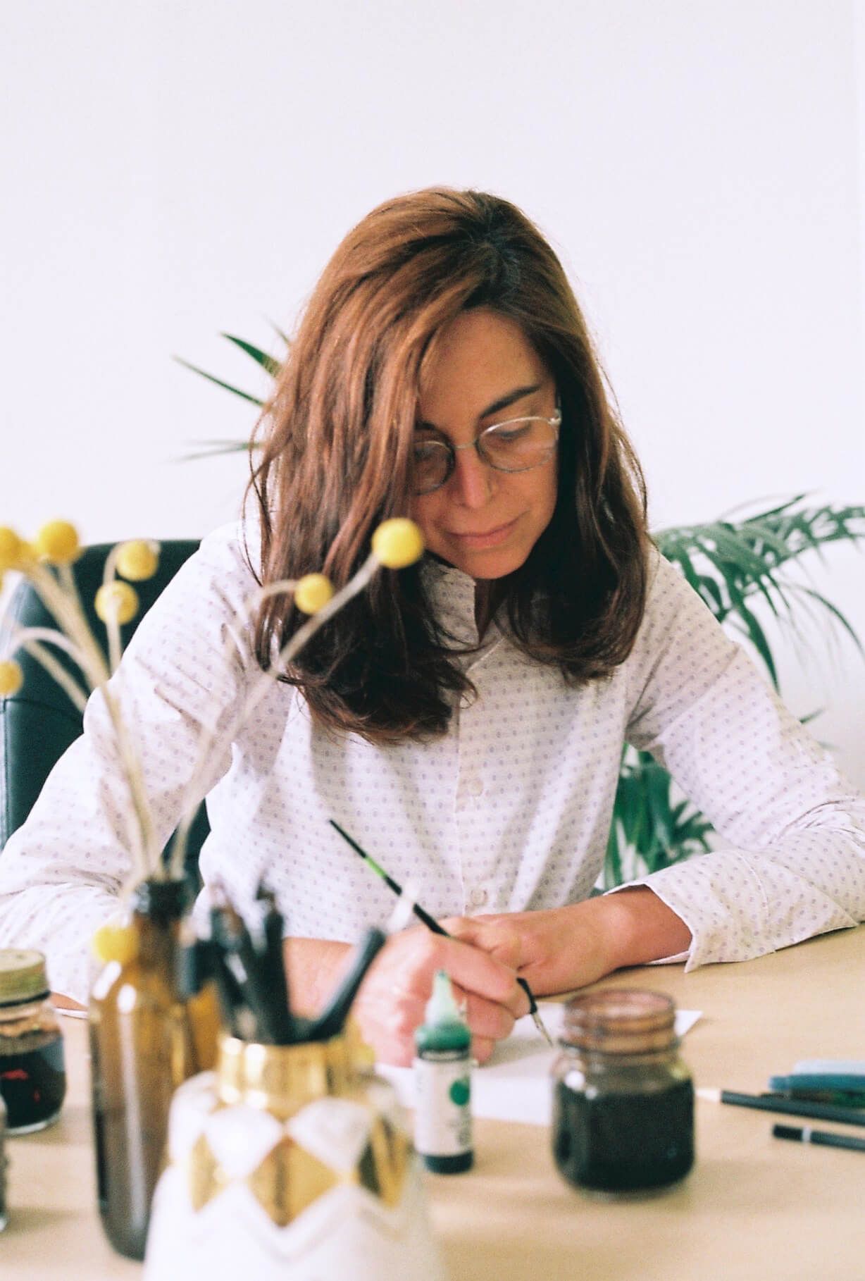 Maria Montes writing at a desk with art supplies and a jar of ink in a bright studio