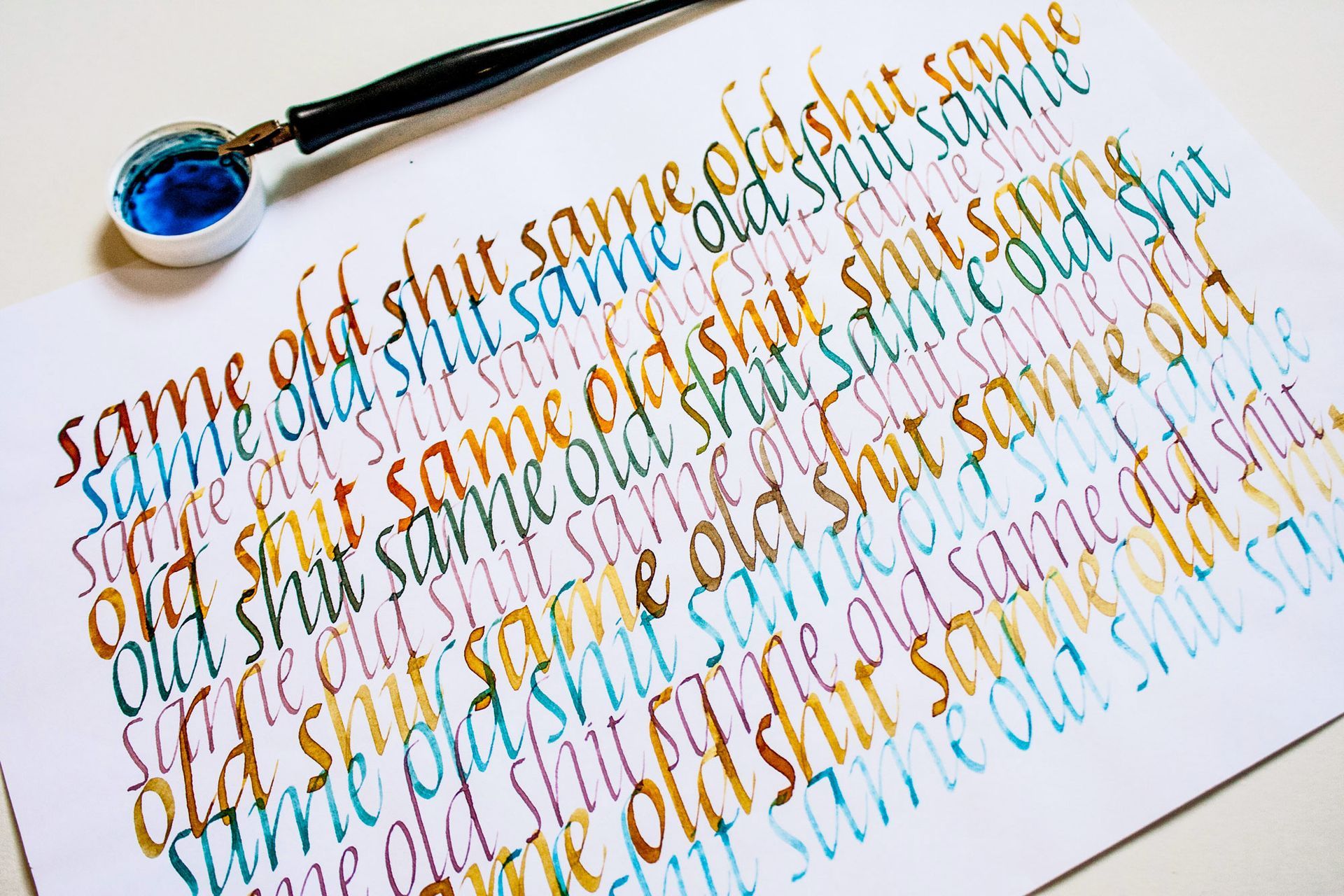 A calligraphy pen rests on paper filled with repetitive, multi-colored script of the phrase 