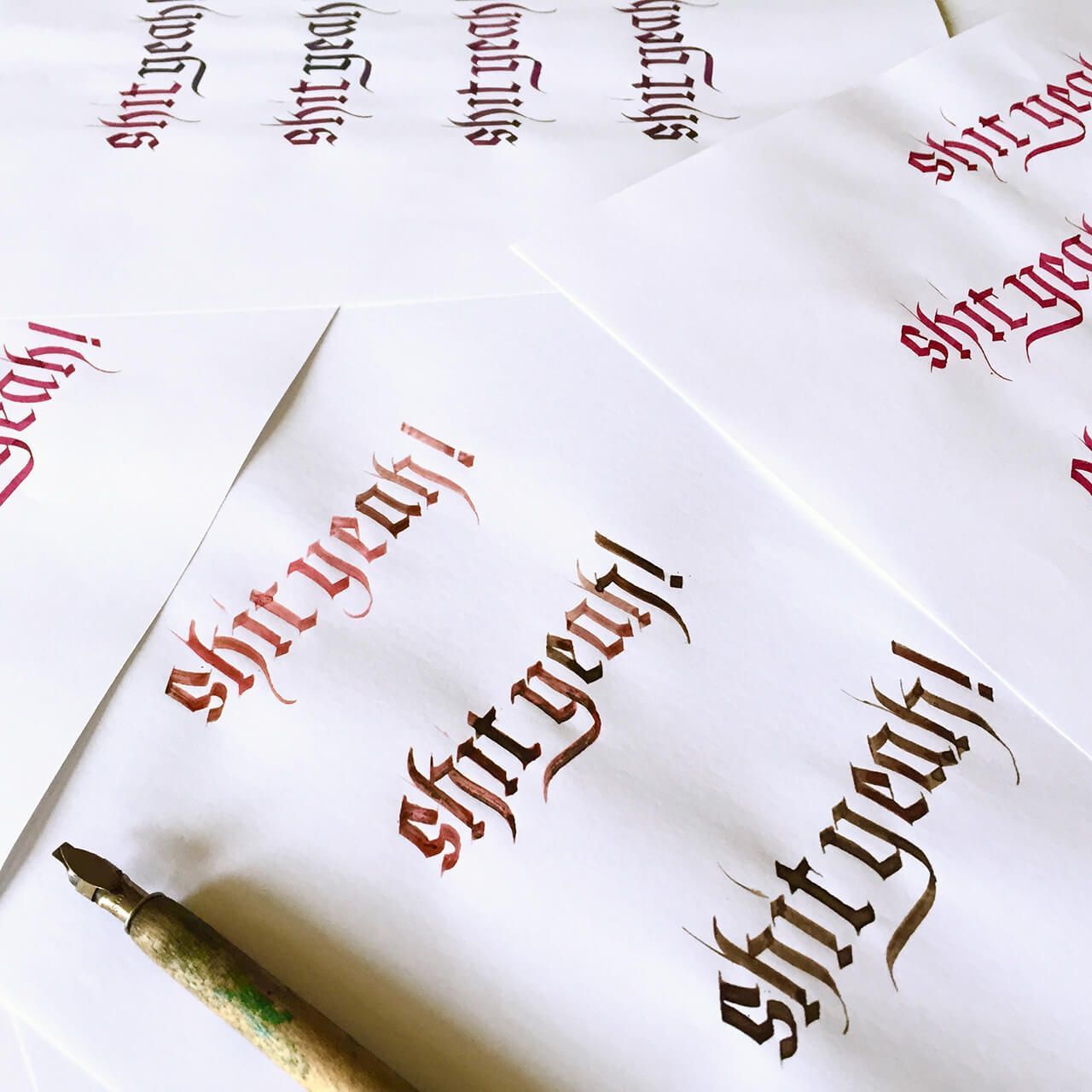 A calligraphy pen lies on white paper covered in the repeated, dark red gothic-style lettering: 