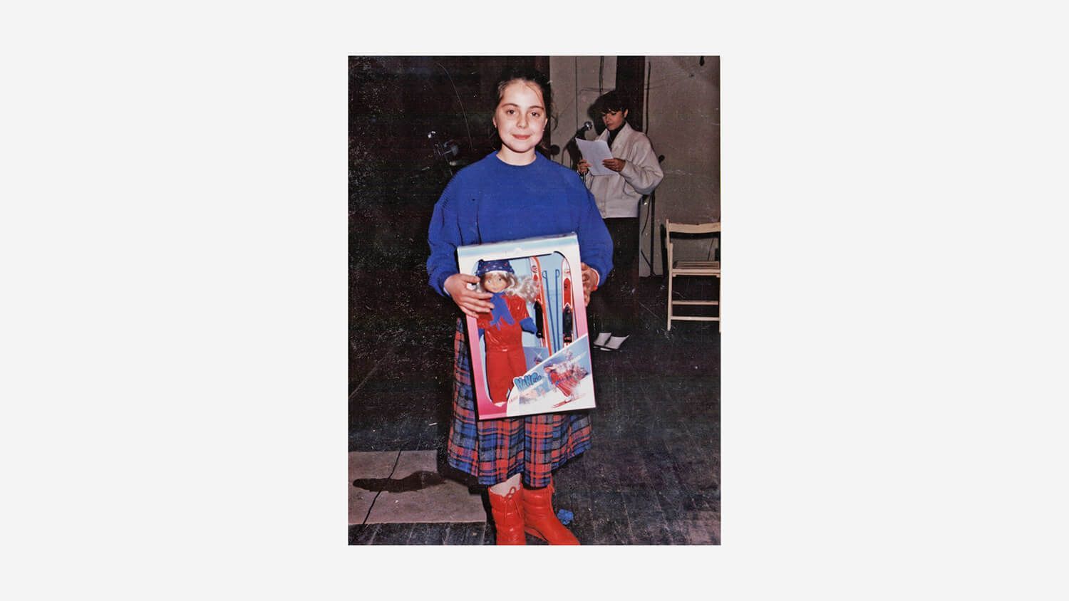 Young Maria Montes in blue top and red plaid pants holds a colorful poster indoors.