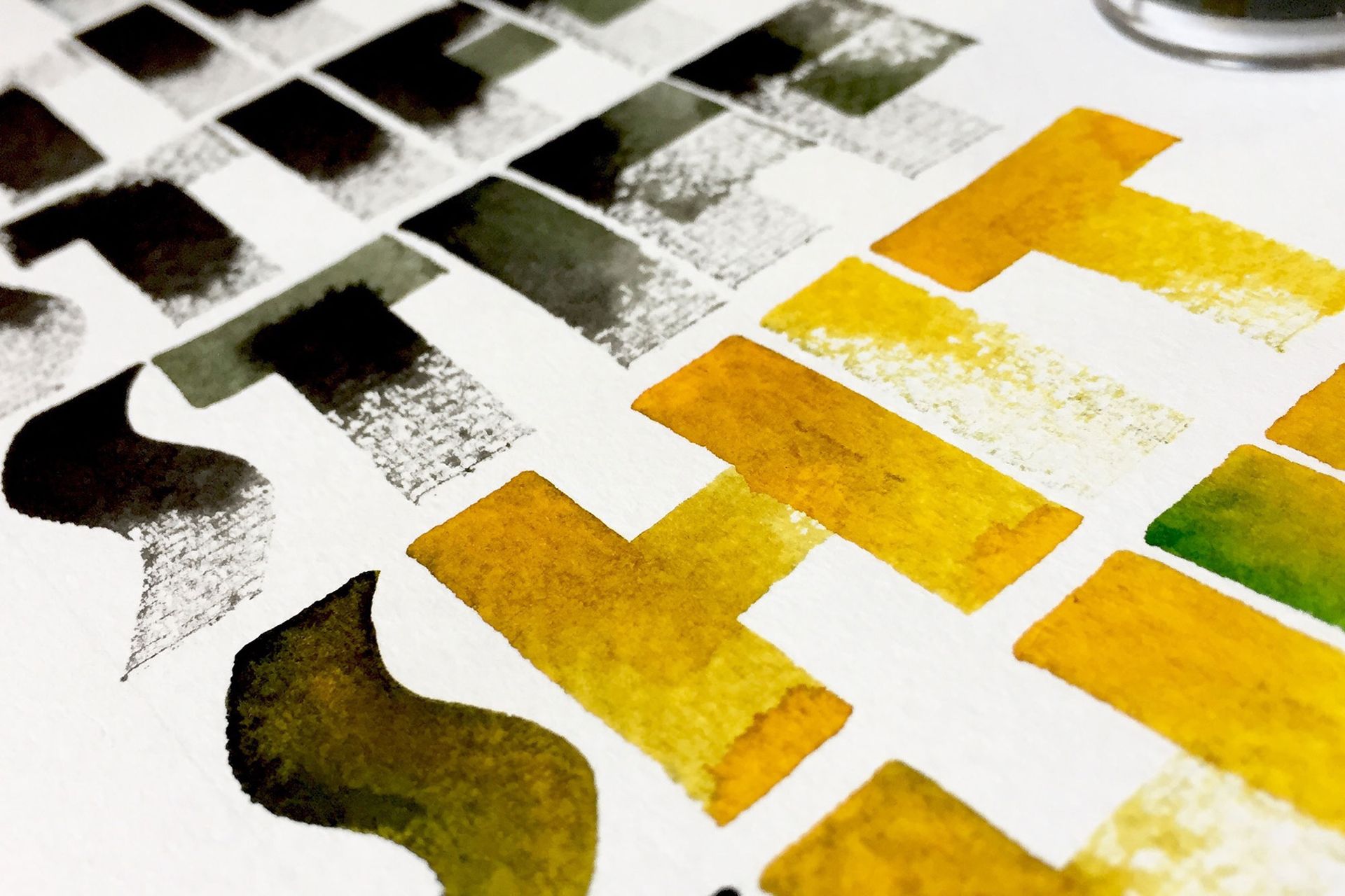A close-up view of watercolor paint swatches in shades of yellow, green, and dark grey on white paper.