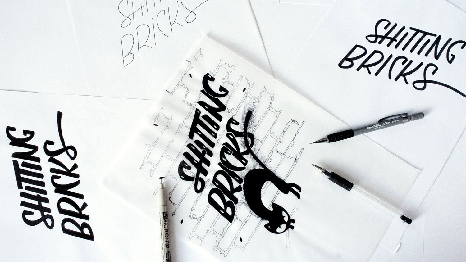 Handwritten “Shitting Bricks” logo sketches on white paper with a pen nearby