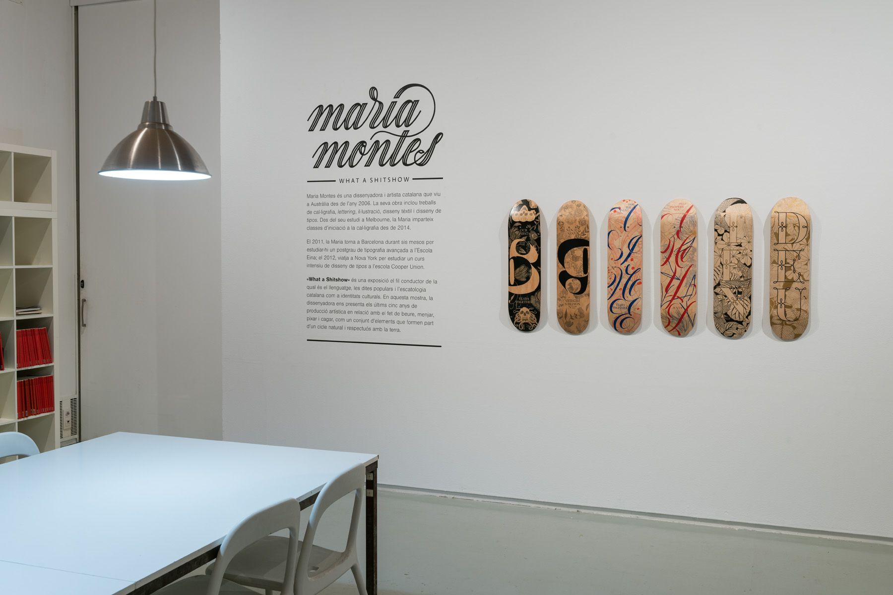 A minimalist room with a table, chairs, a hanging lamp, and six decorated skateboards mounted on a wall beside text.