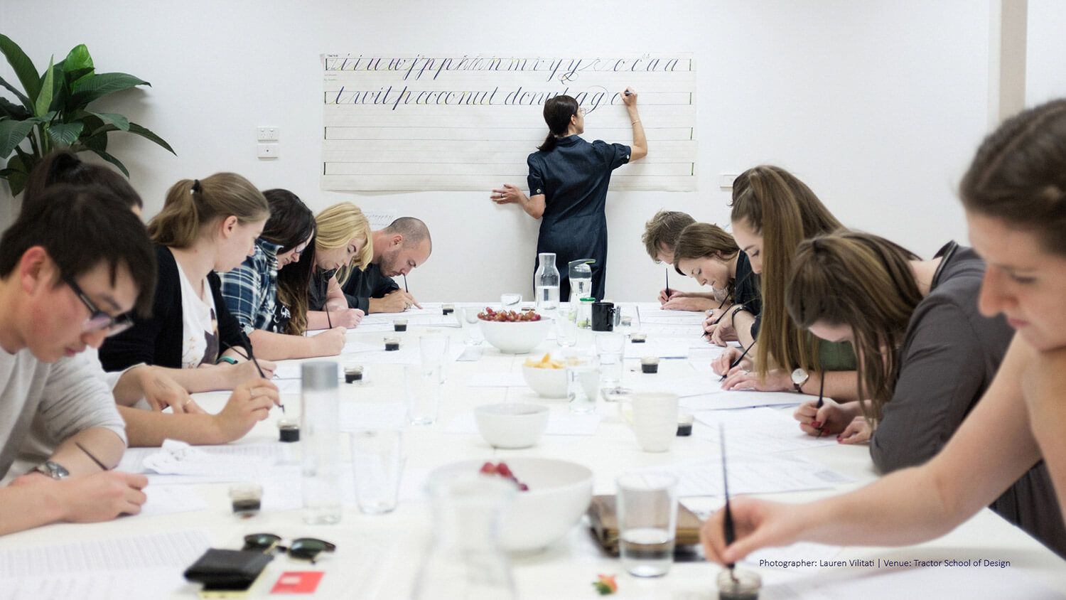 People seated at a long table in a bright classroom, taking notes while a presenter writes on a whiteboard.