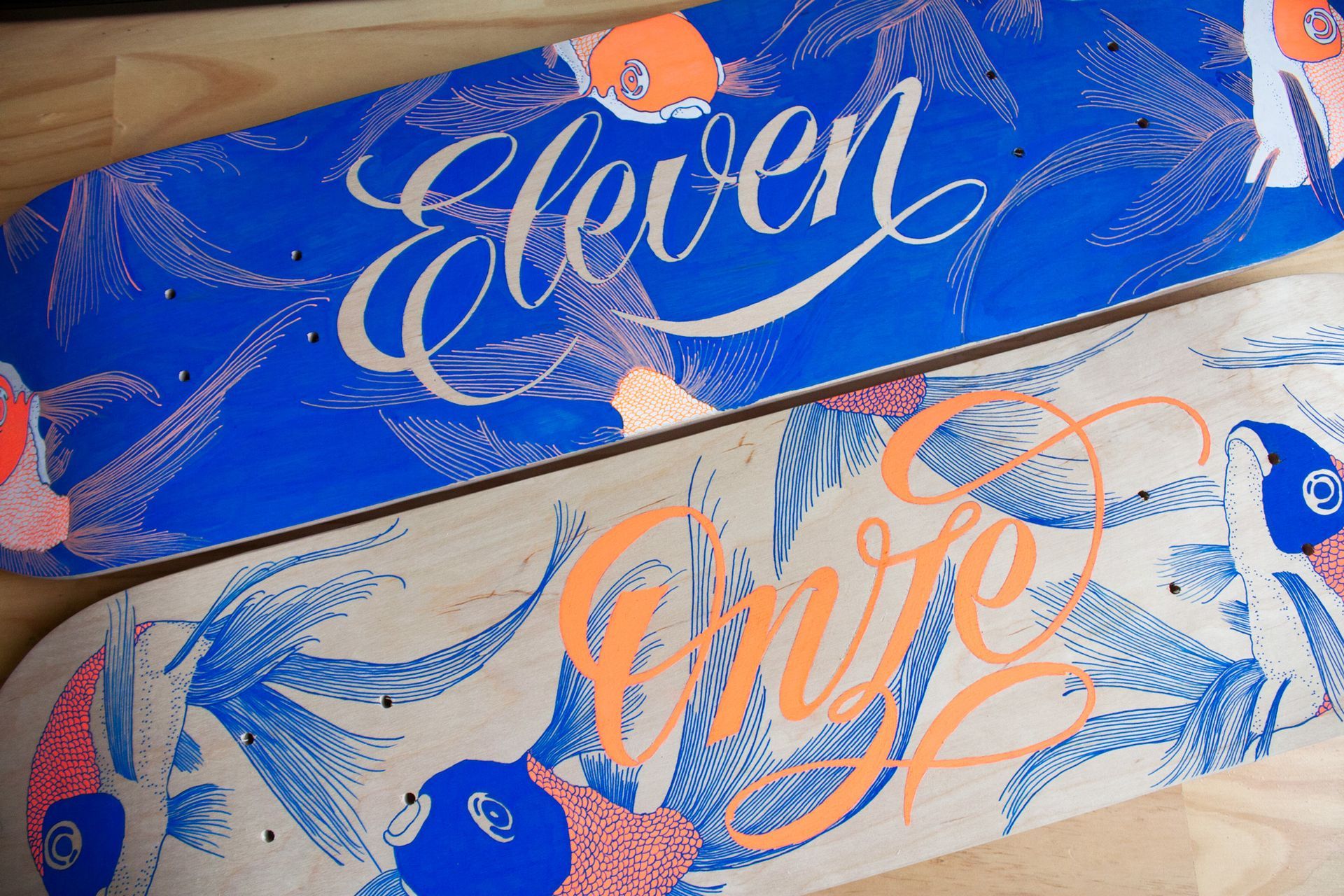 Two skateboards with matching blue and orange stylized fish patterns; one has a blue background and one a natural wood.