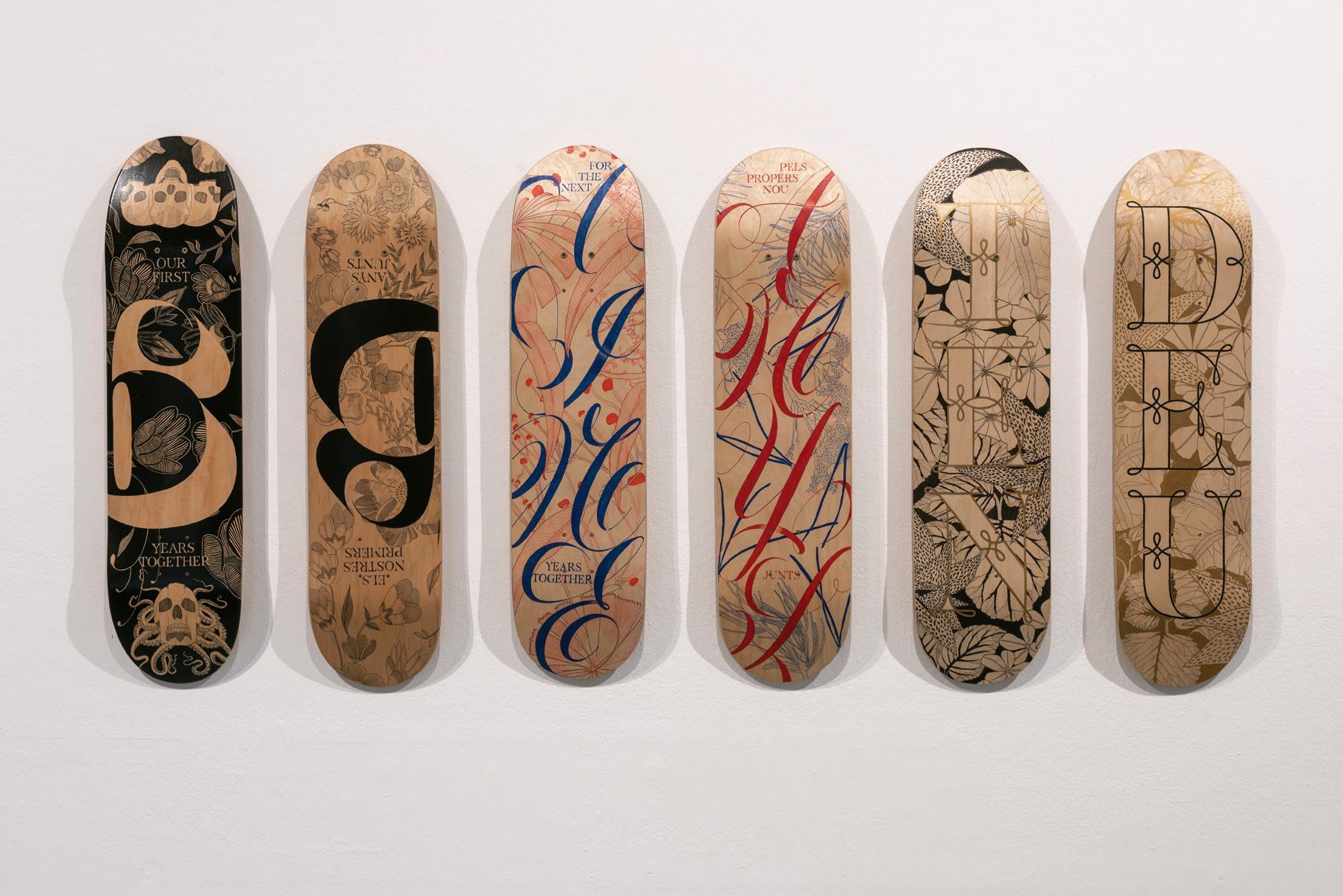 Six skateboards with diverse typographic and decorative art designs mounted in a row against a plain white wall.