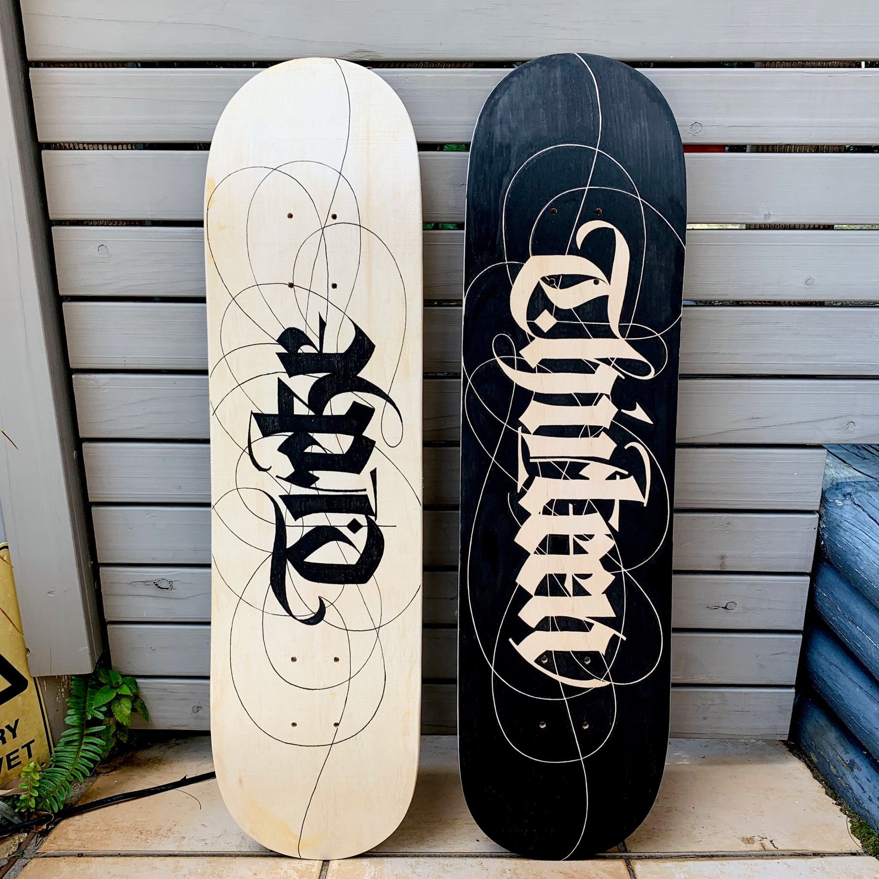 Two skateboards leaning against a gray wooden wall, one light wood with black lettering and the other black with white text.