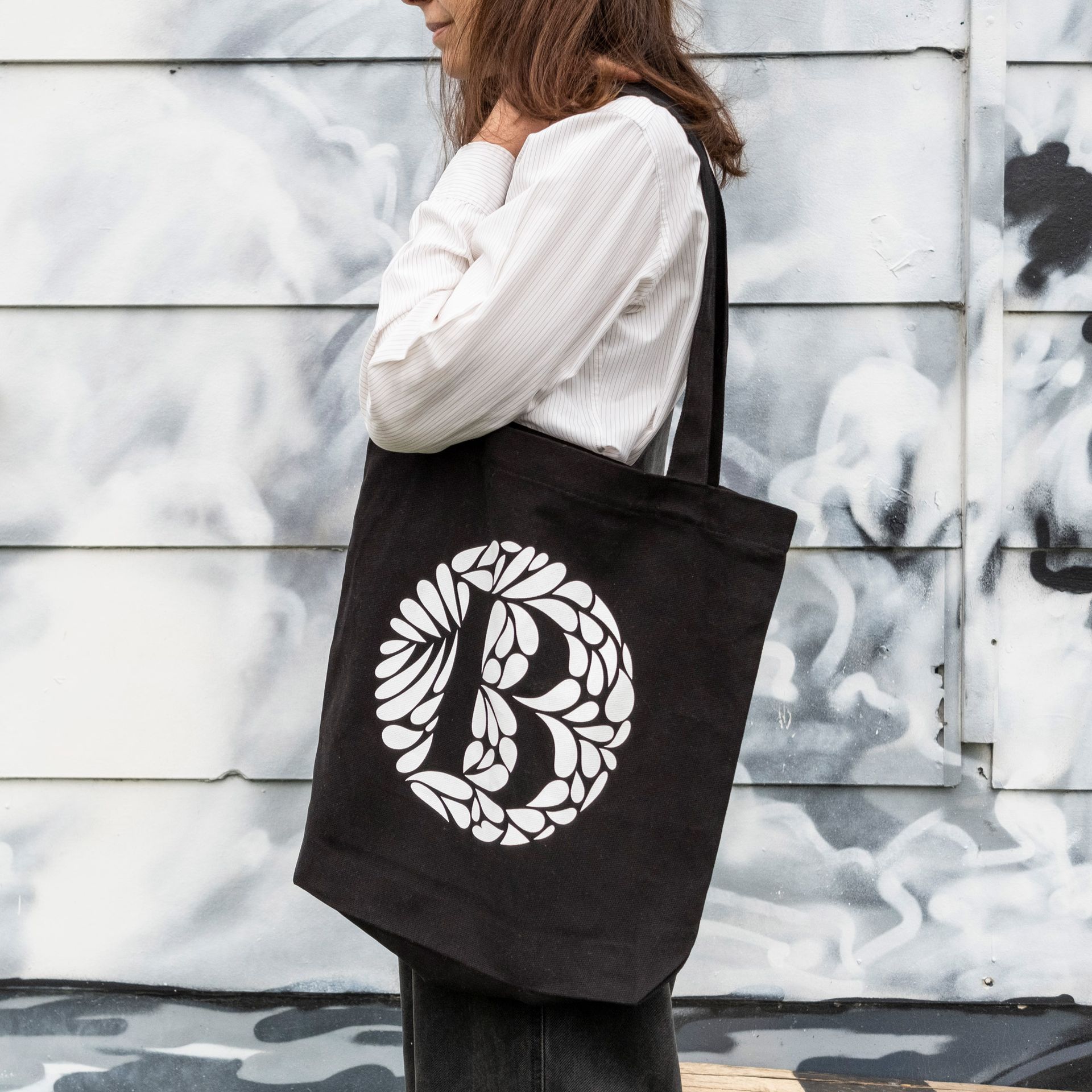 Person carrying a black tote bag with a white floral letter B design, standing by a graffiti wall
