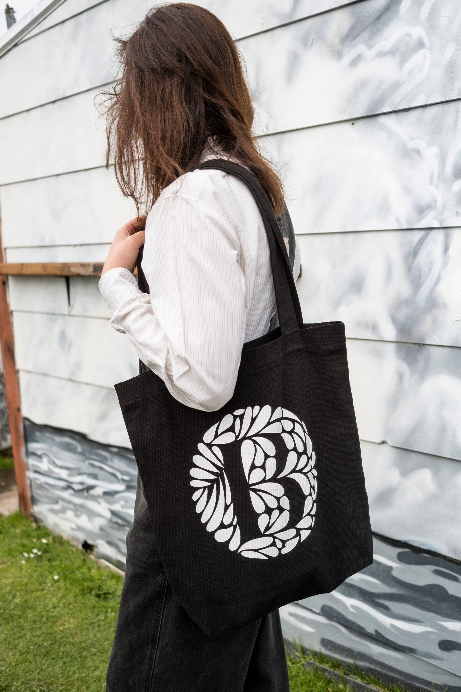 Maria Montes in a white shirt and black overalls carrying a black tote bag with a white design, standing by a wall.
