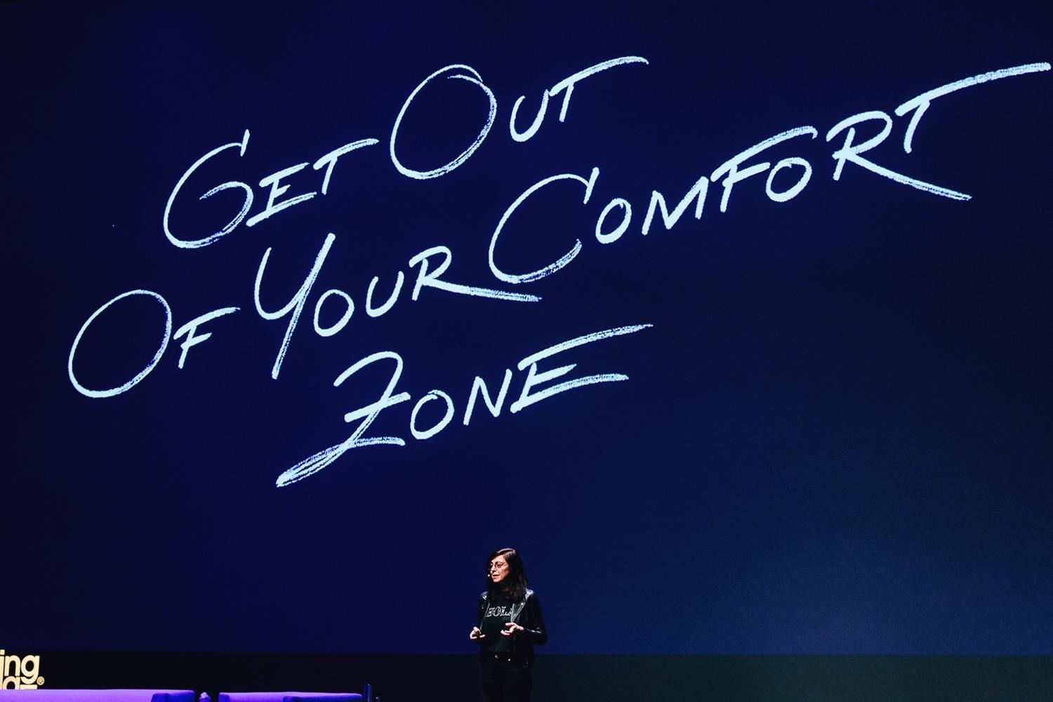 Large stage screen with handwritten text, “Get Out Of Your Comfort Zone,” above Maria Montes on stage