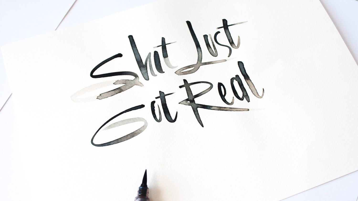 Handwritten “Shut Just Sit Real” in black ink on white paper, with a pen tip at the bottom edge