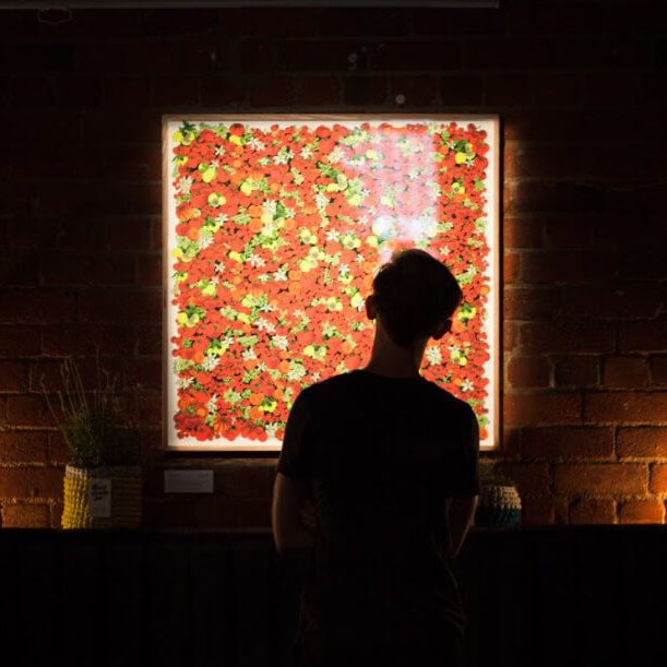 Silhouette of a person facing a brightly lit floral artwork in a dark brick-walled gallery
