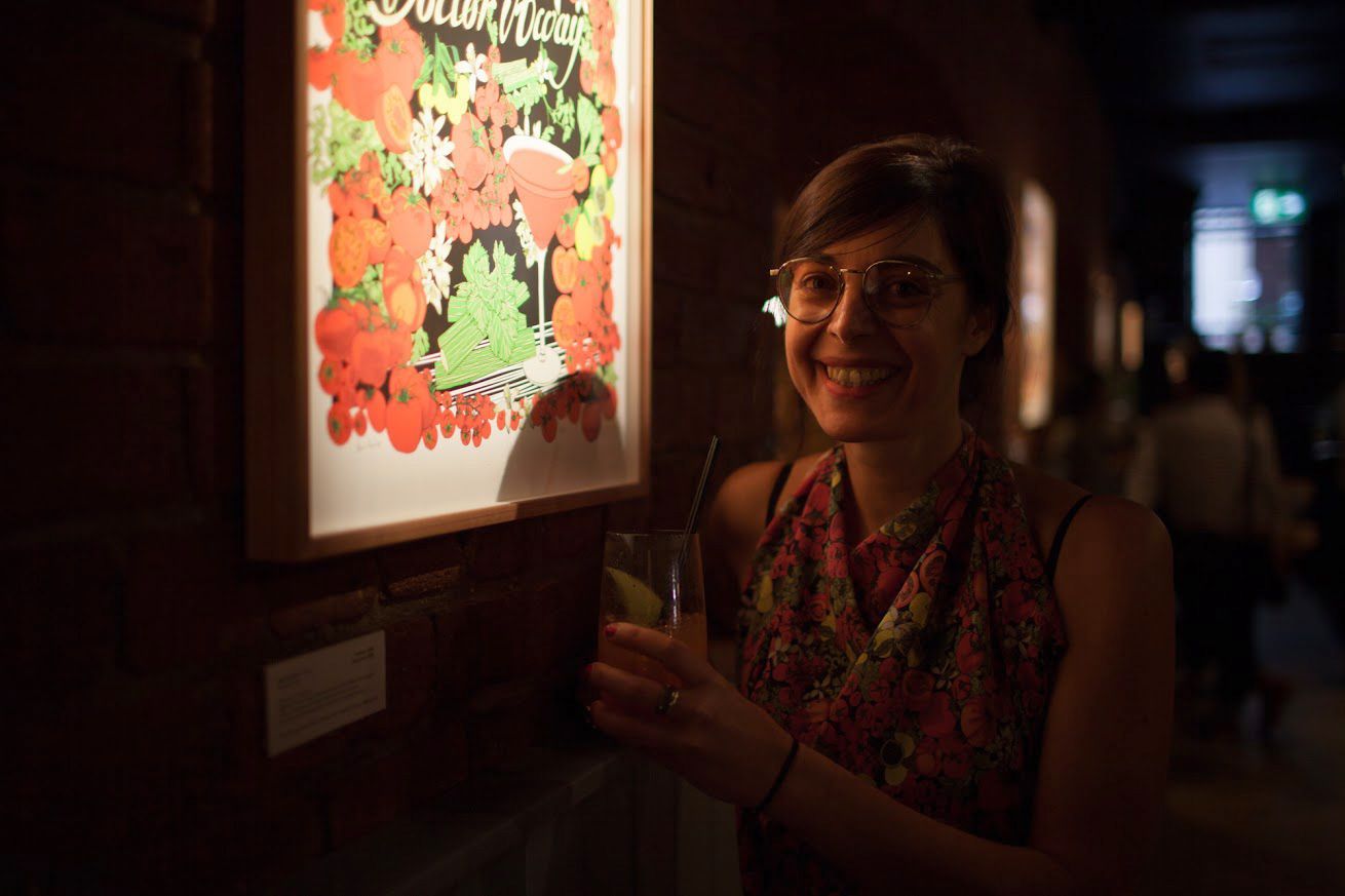 Maria Montes smiling beside a brightly lit floral artwork in a dark gallery hallway
