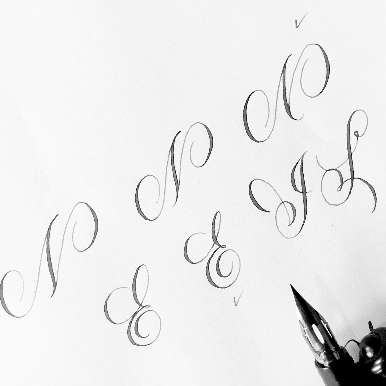 A calligraphy pen rests on paper beside several elegant, hand-lettered cursive capital letters.