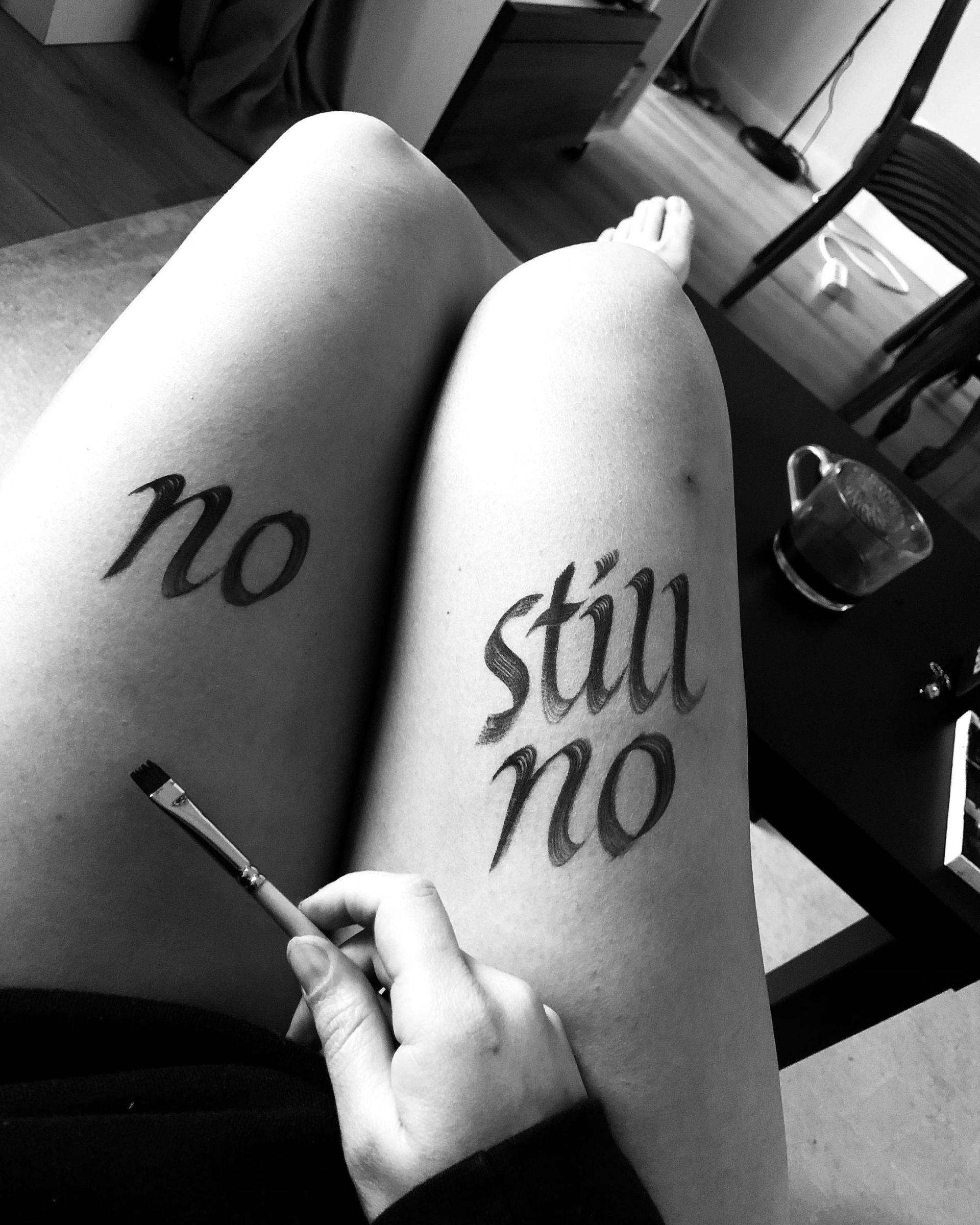 Black-and-white photo of legs with “no” and “still no” written on them, with a cigarette in hand.