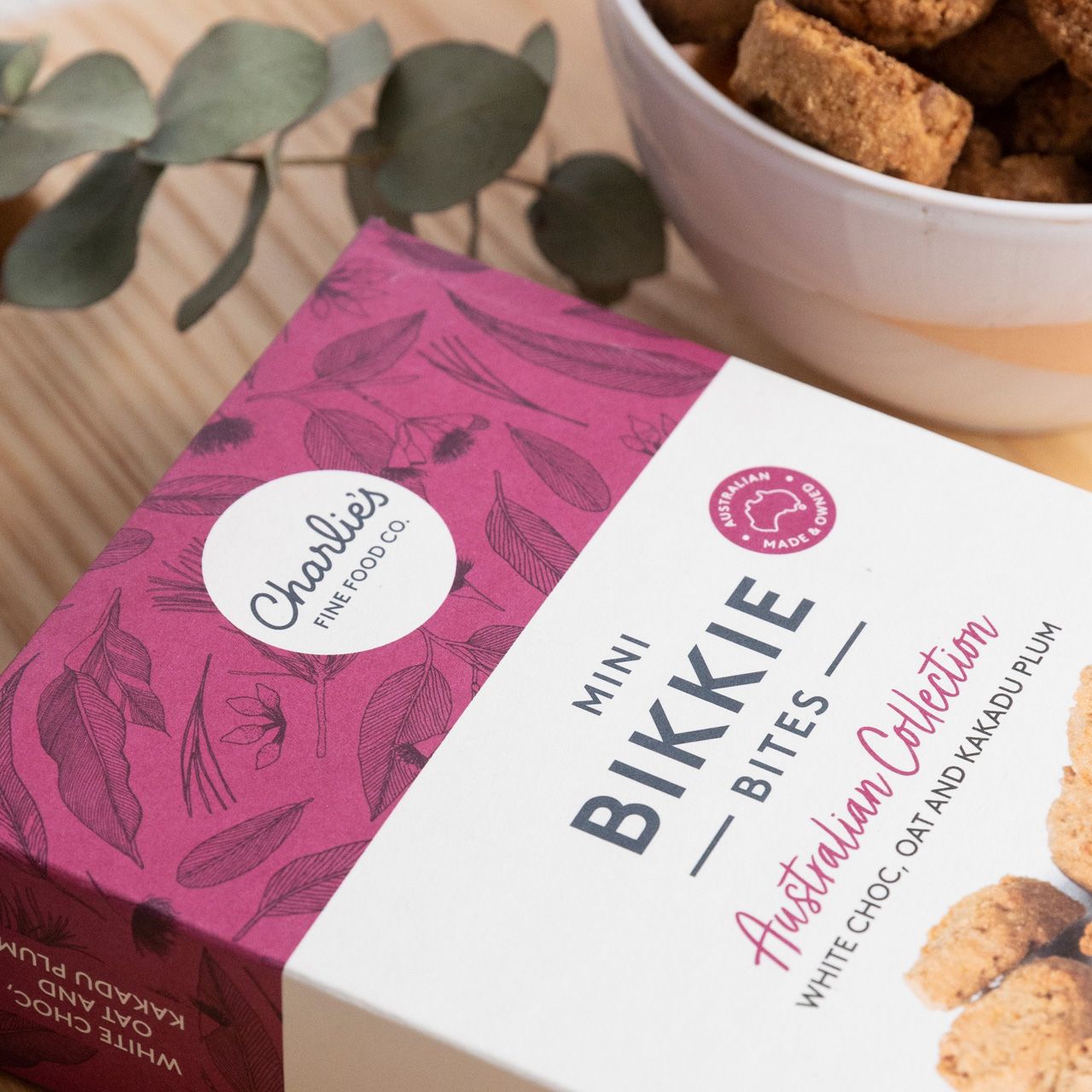 Pink-and-white box of mini bikkie bites with coconut flakes and a bowl of biscuits on a wooden table